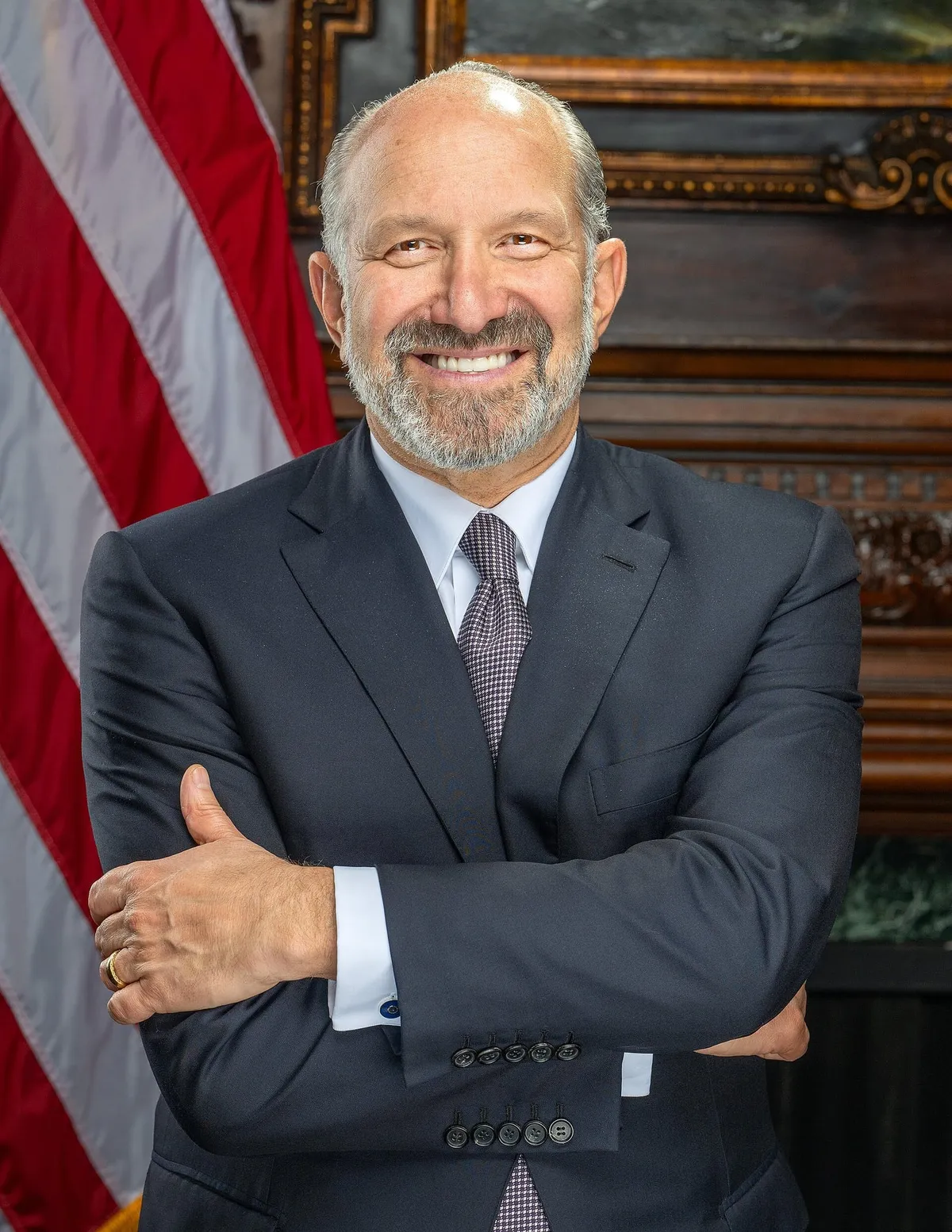 Official portrait of Commerce Secretary Howard Lutnick, arms crossed, beside a U.S. flag