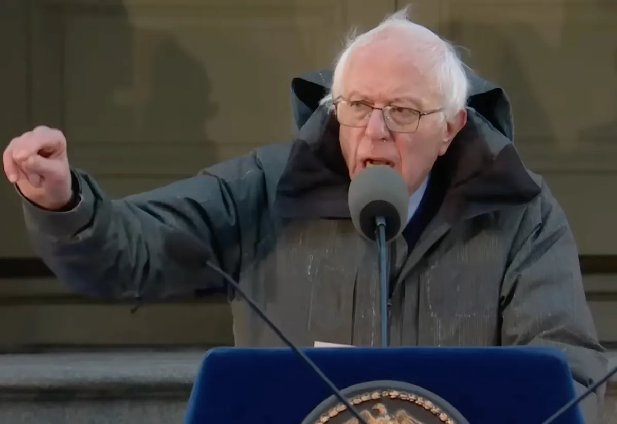 Bernie Sanders speaking at a lectern outdoors in a winter coat, fist raised
